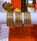 Sets of gold-plated bangles with intricate floral and geometric engravings, displayed on a white cylindrical stand. The background features a branded orange and white SAWRA GOLD box.