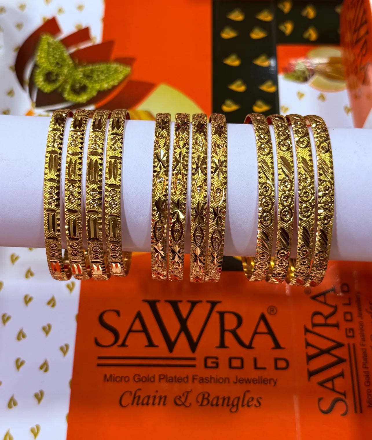 A set of ornate gold-plated bangles displayed on a white cylindrical stand, with SAWRA GOLD branding visible on the packaging in the background.