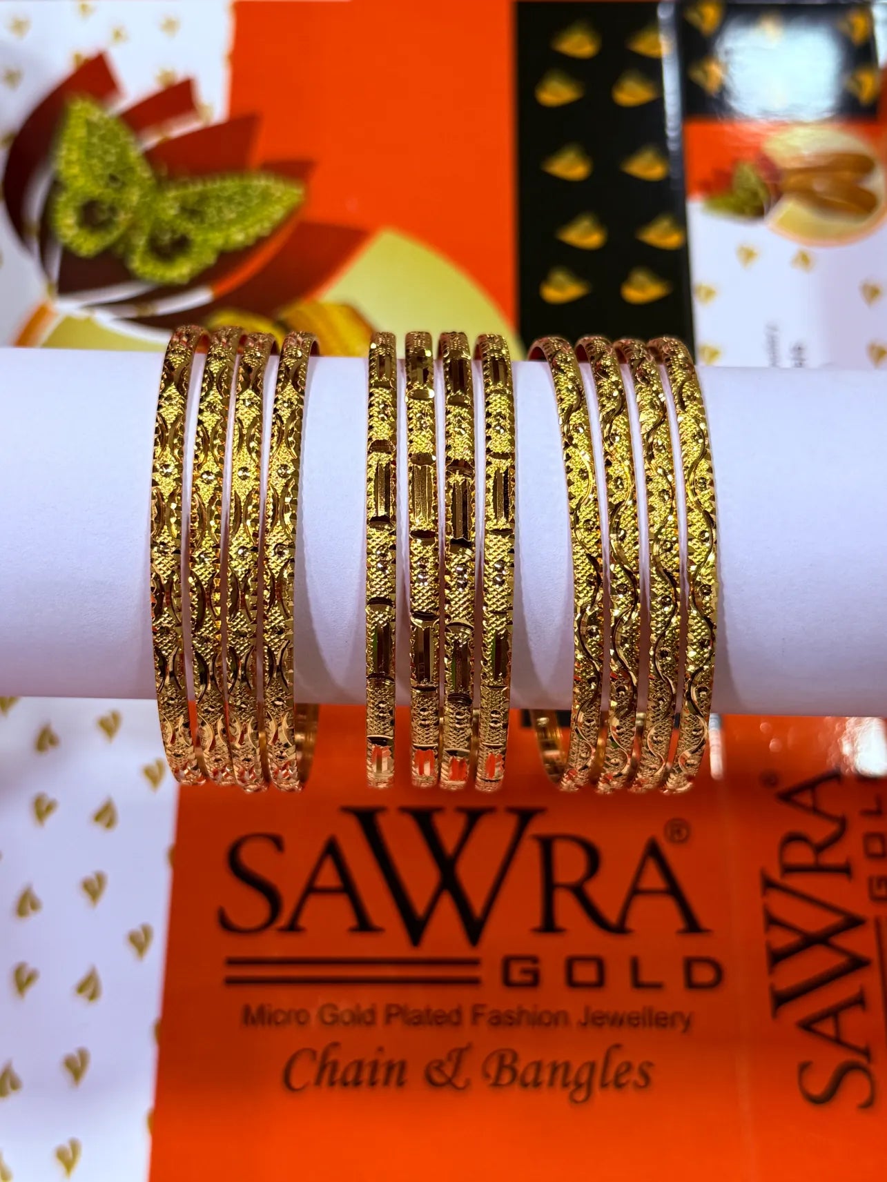 A set of gold-plated bangles displayed on a white cylindrical stand, with SAWRA GOLD branding visible in the background packaging.